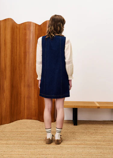 Person wearing a navy pinafore dress with a white shirt underneath, standing in a room with wooden paneling and a whiteboard.